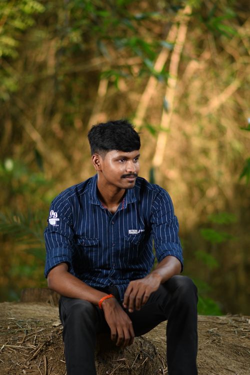 Free Young man sitting outdoors in nature, wearing a blue striped shirt, looking thoughtful. Stock Photo