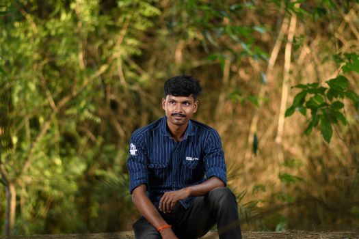 A man sitting outdoors against a lush green background, wearing a striped shirt.
