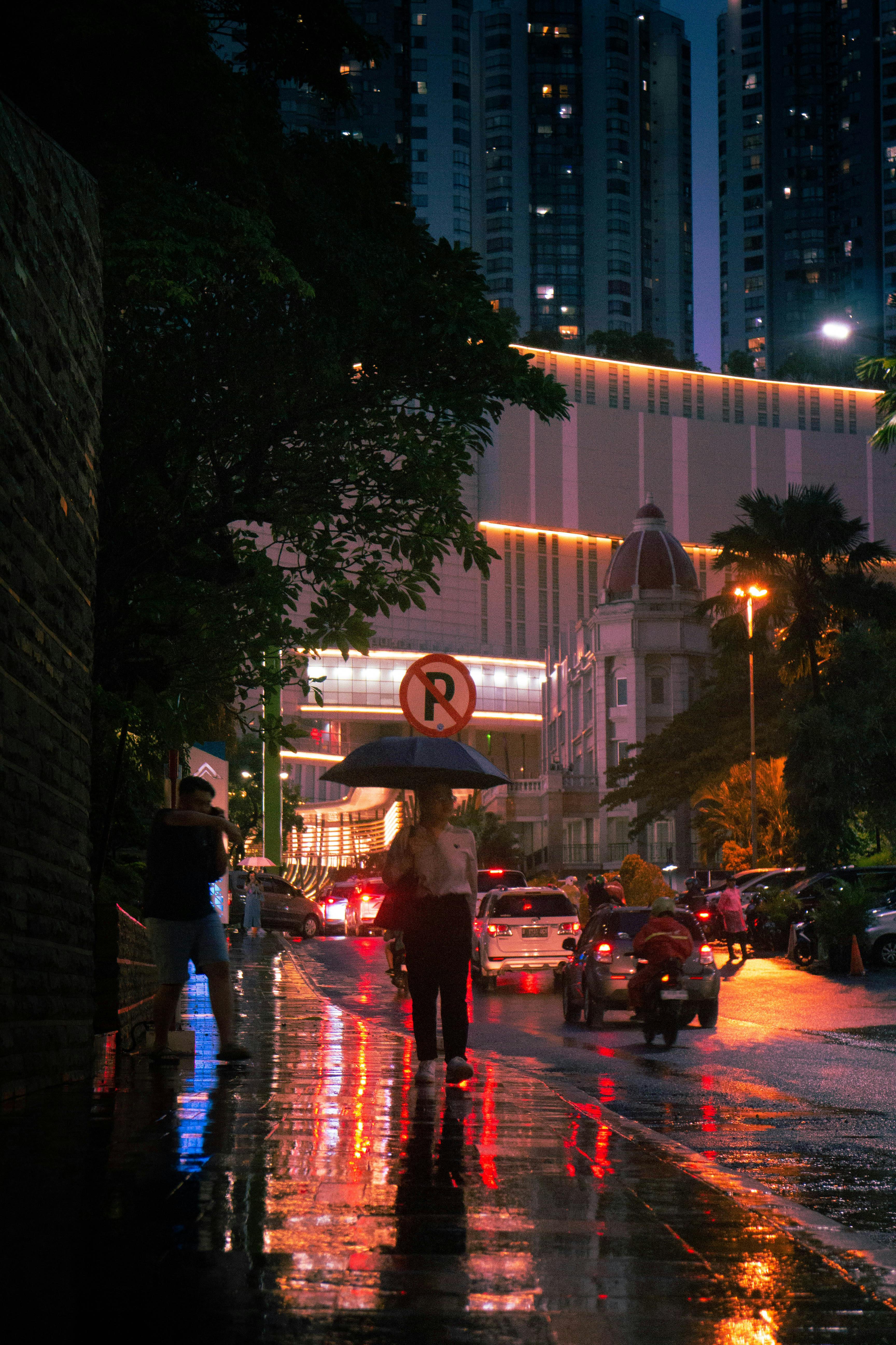 A person with an umbrella walks down a wet street at night, surrounded by tall buildings and city lights.