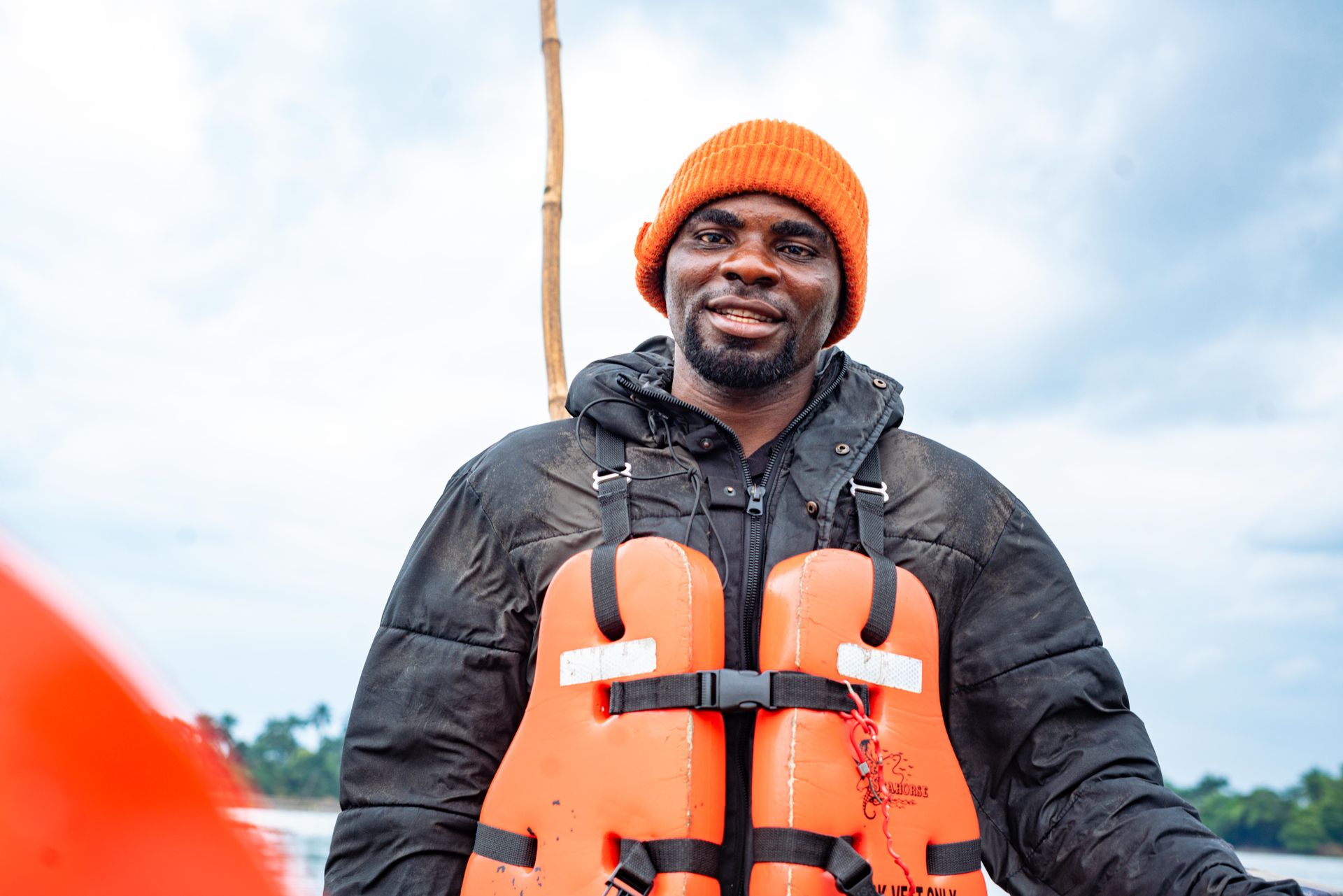 A man wearing a life jacket with an orange beanie smiles confidently outdoors.
