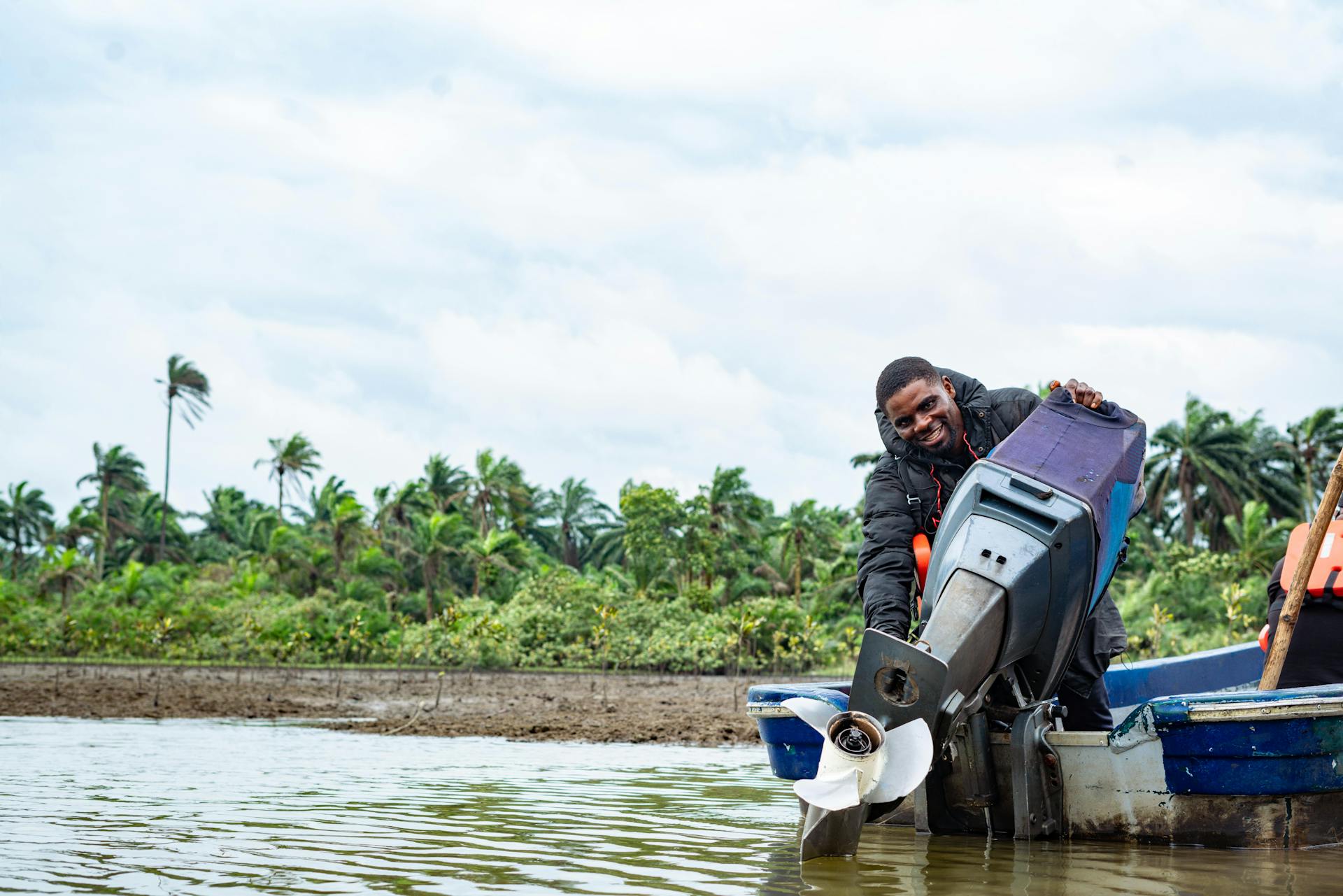A man repairs a boat engine near tropical vegetation on a sunny day.