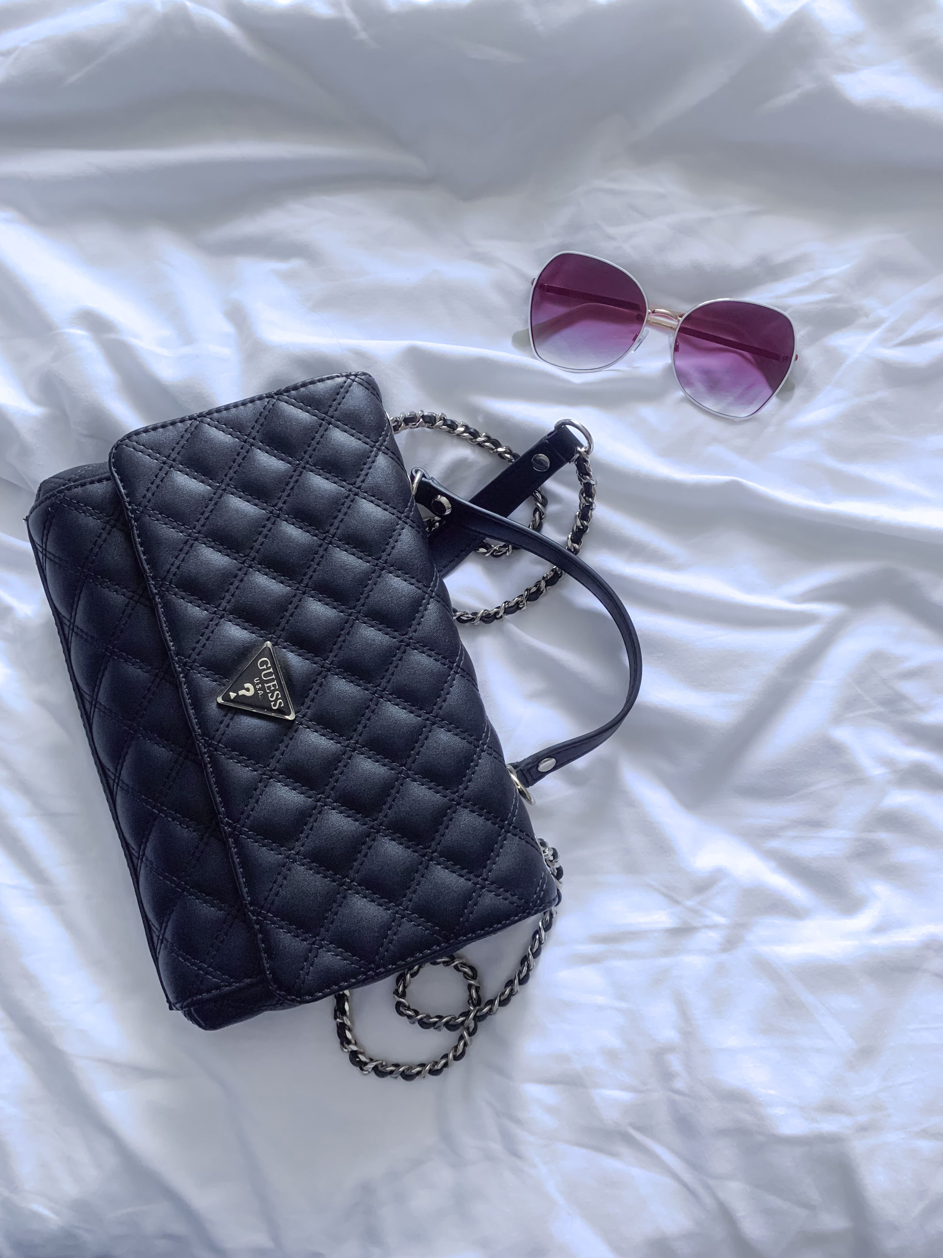 Bolsa de mão preta acolchoada e óculos de sol dispostos na cama branca para um estilo elegante.