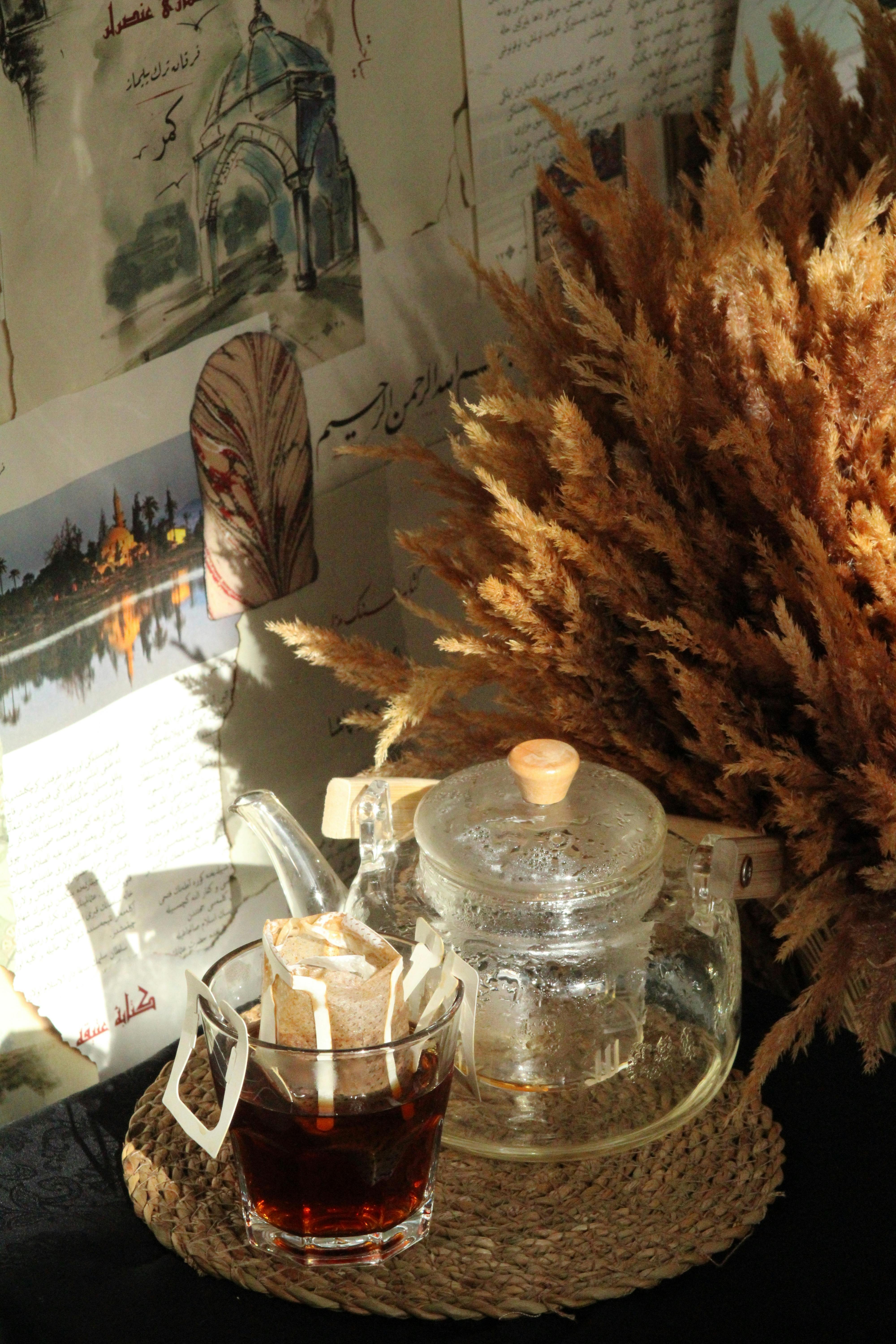 A clear glass teapot and cup of tea with a dried floral arrangement in warm lighting.