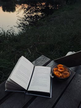 An open book on a bench with a bowl of apricots beside a tranquil lake at sunset.