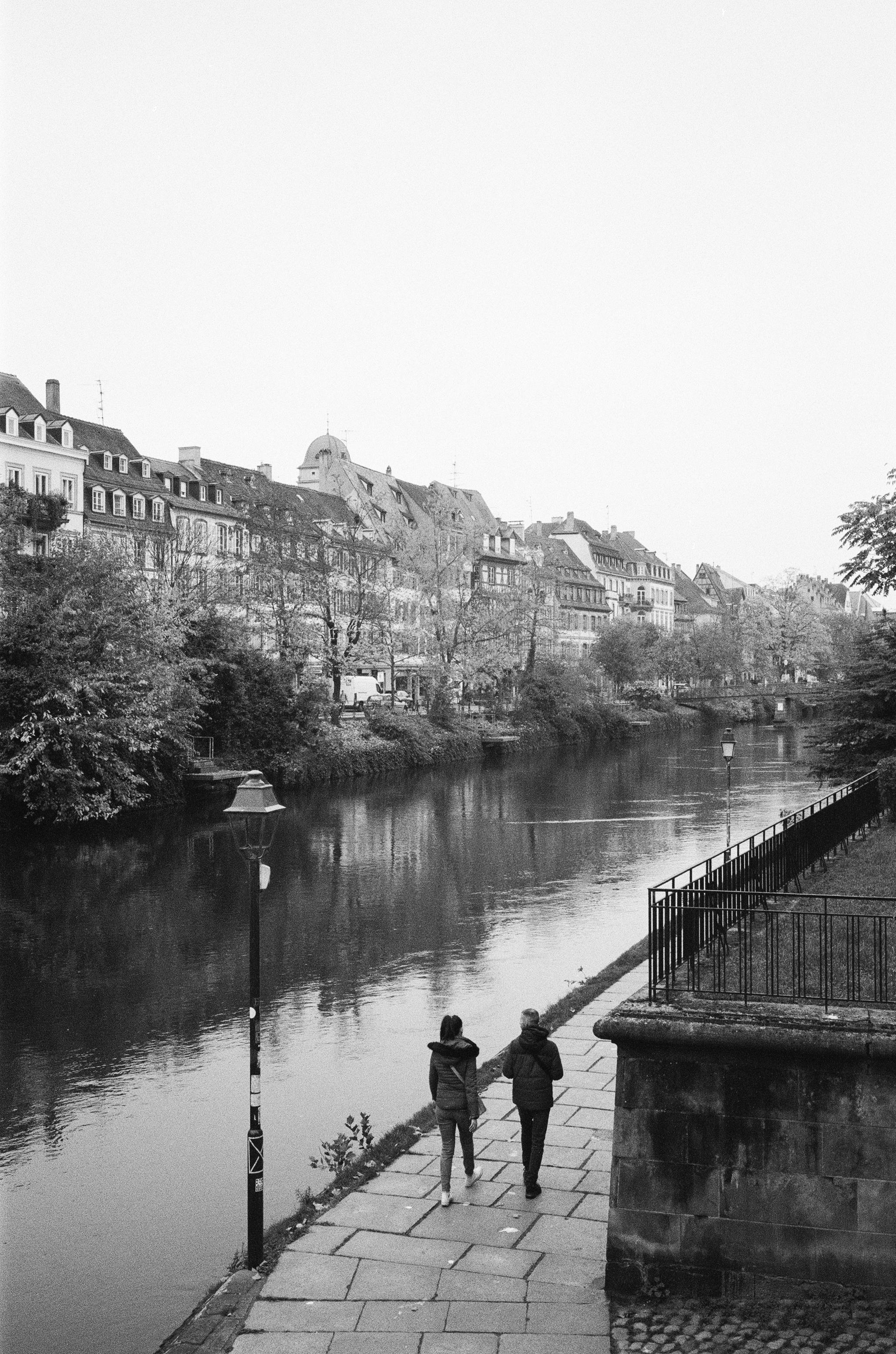 Black and white photograph of a riverside walk in Strasbourg, capturing historic architecture and serene atmosphere.