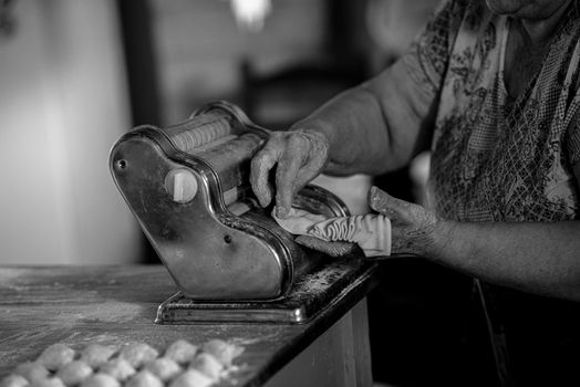 An elderly woman uses a pasta machine to prepare homemade pasta, captured in black and white.