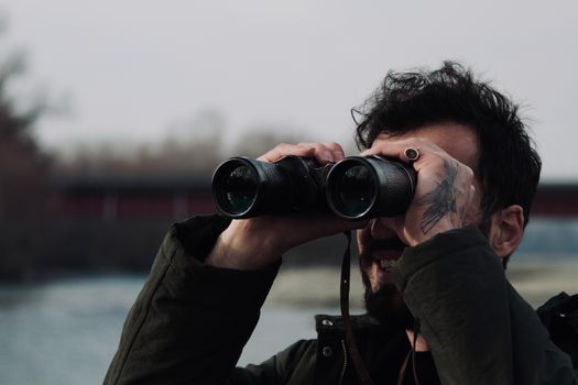 A man with a tattoo observes with binoculars near a river, showcasing exploration.