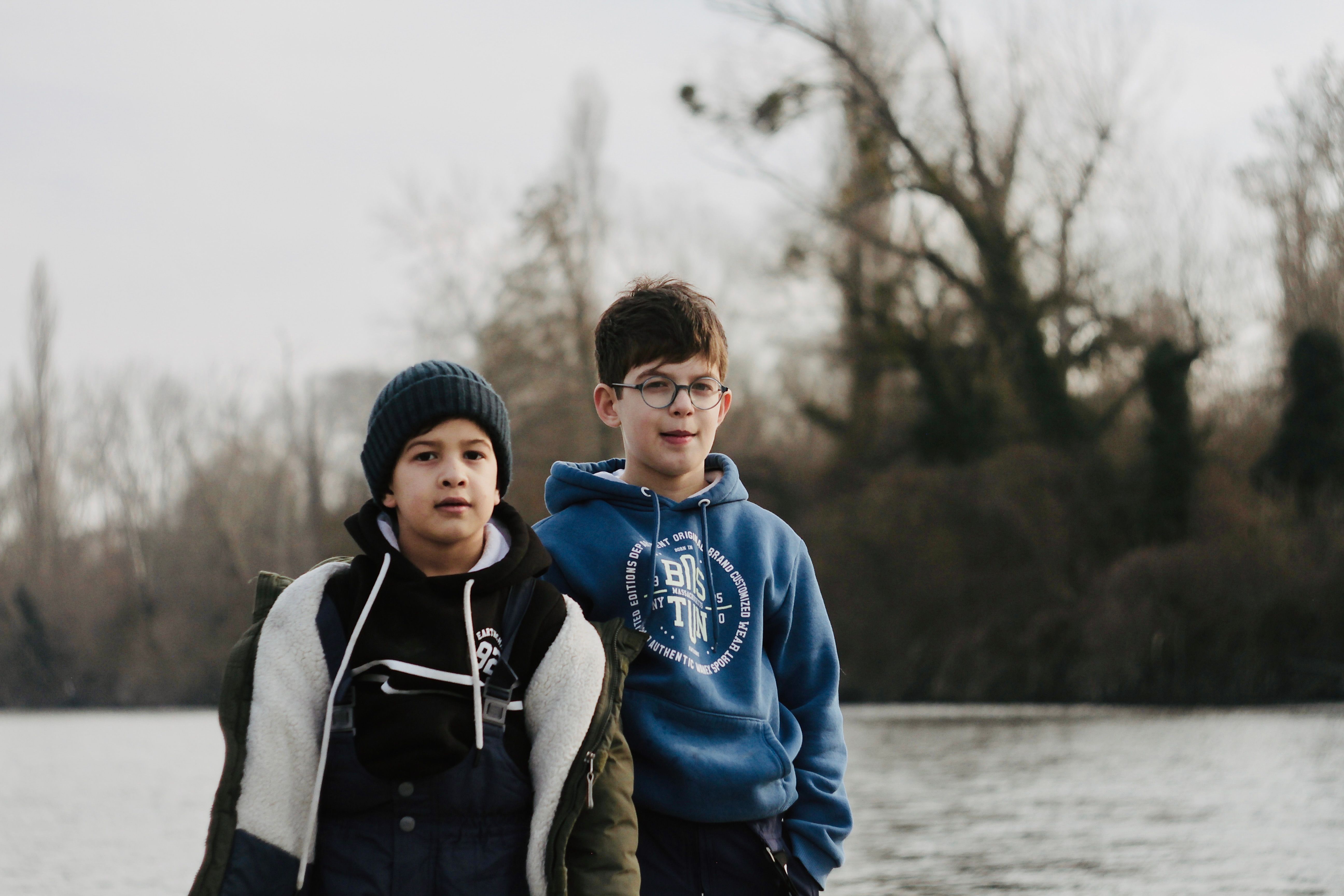 Two young boys in winter apparel stand by a serene lake with trees.