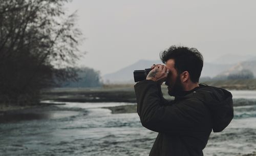 Free Man with binoculars observing nature by a scenic river in a tranquil outdoor setting. Stock Photo
