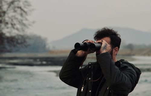 Free A man with tattoos uses binoculars to view a scenic landscape outdoors during the day. Stock Photo