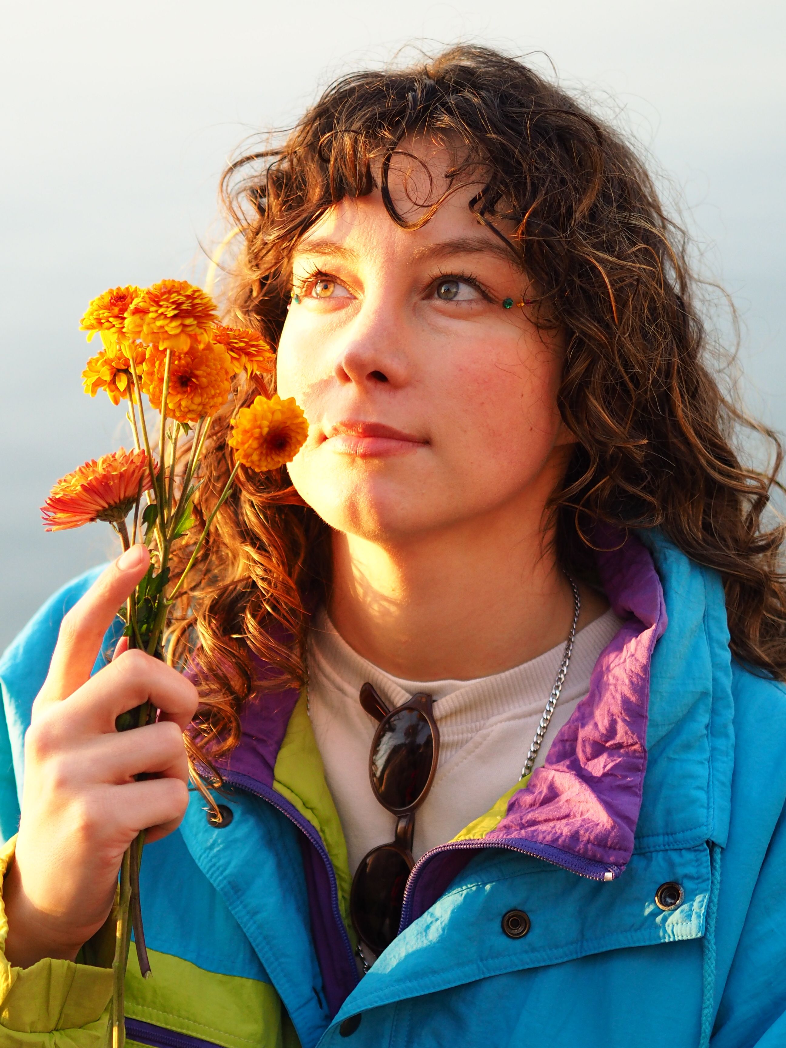A woman gazes upward while holding bright orange flowers, wearing a colorful jacket.