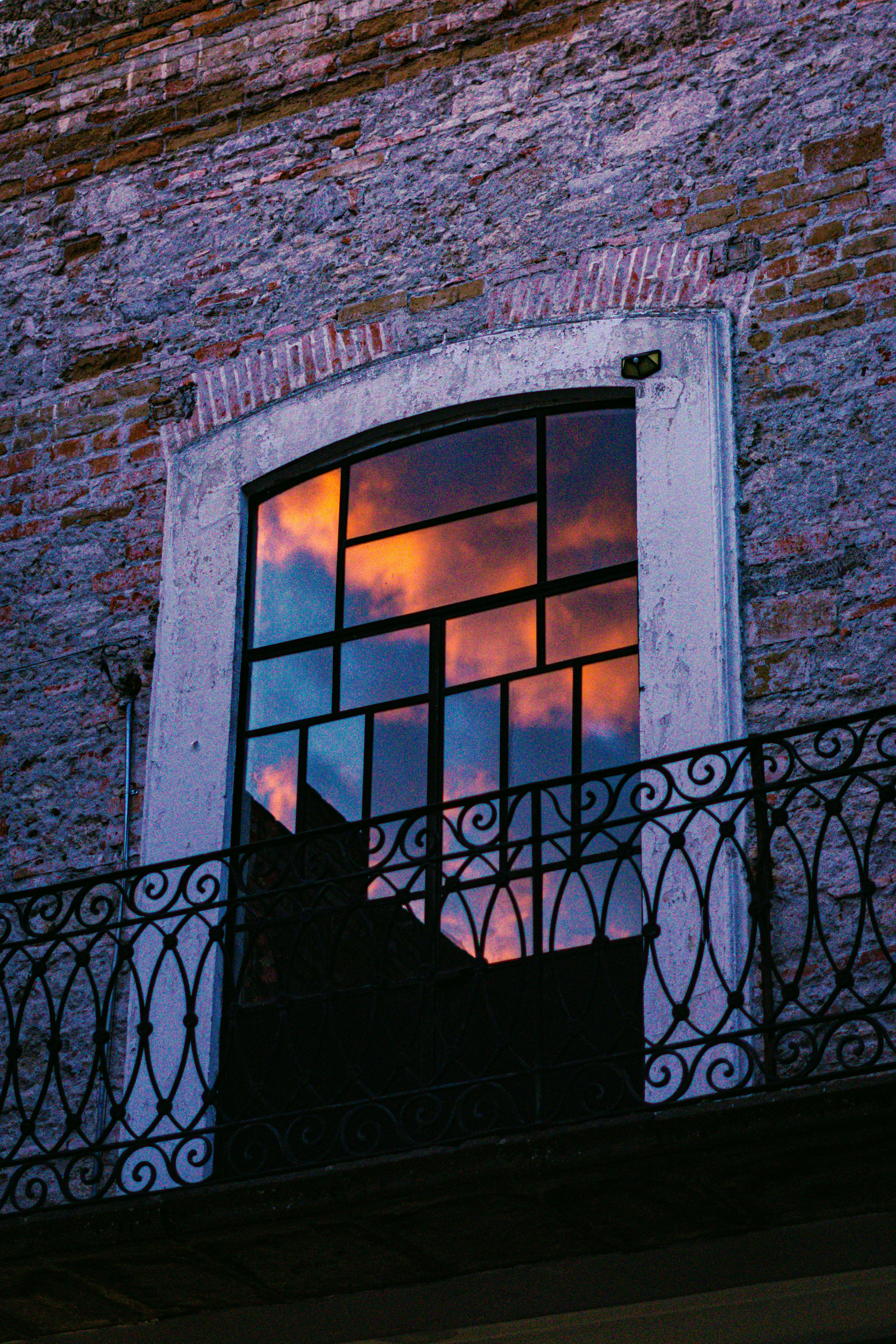 A vivid sunset reflects in an arched window with wrought iron balcony.