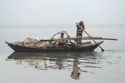 Free Two men on a traditional wooden fishing boat navigating a calm river with oars. Stock Photo