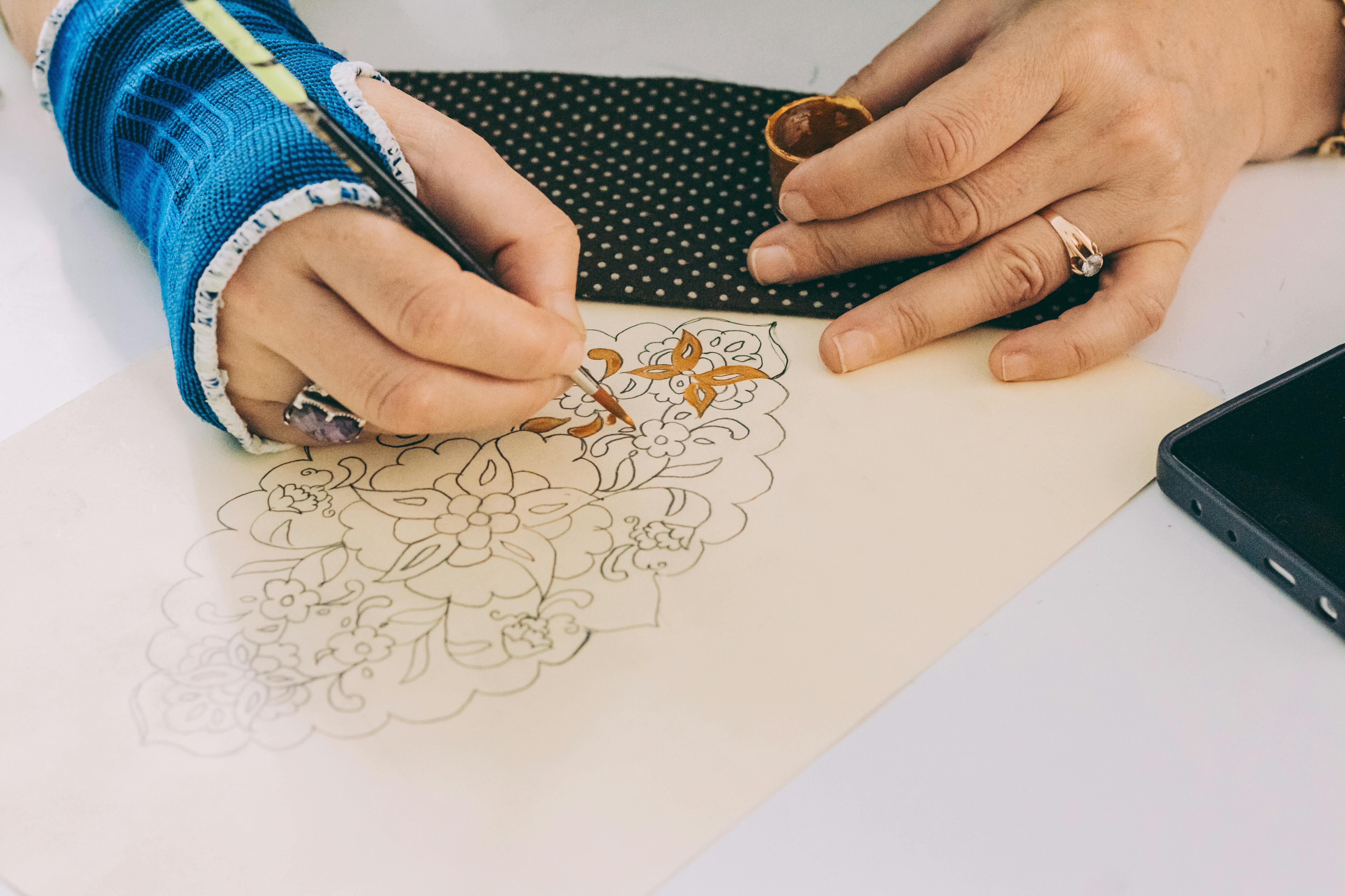 grátis Pintura decorativa à mão em tecido, destacando um intrincado desenho floral com um artista usando um gesso no pulso. Foto profissional