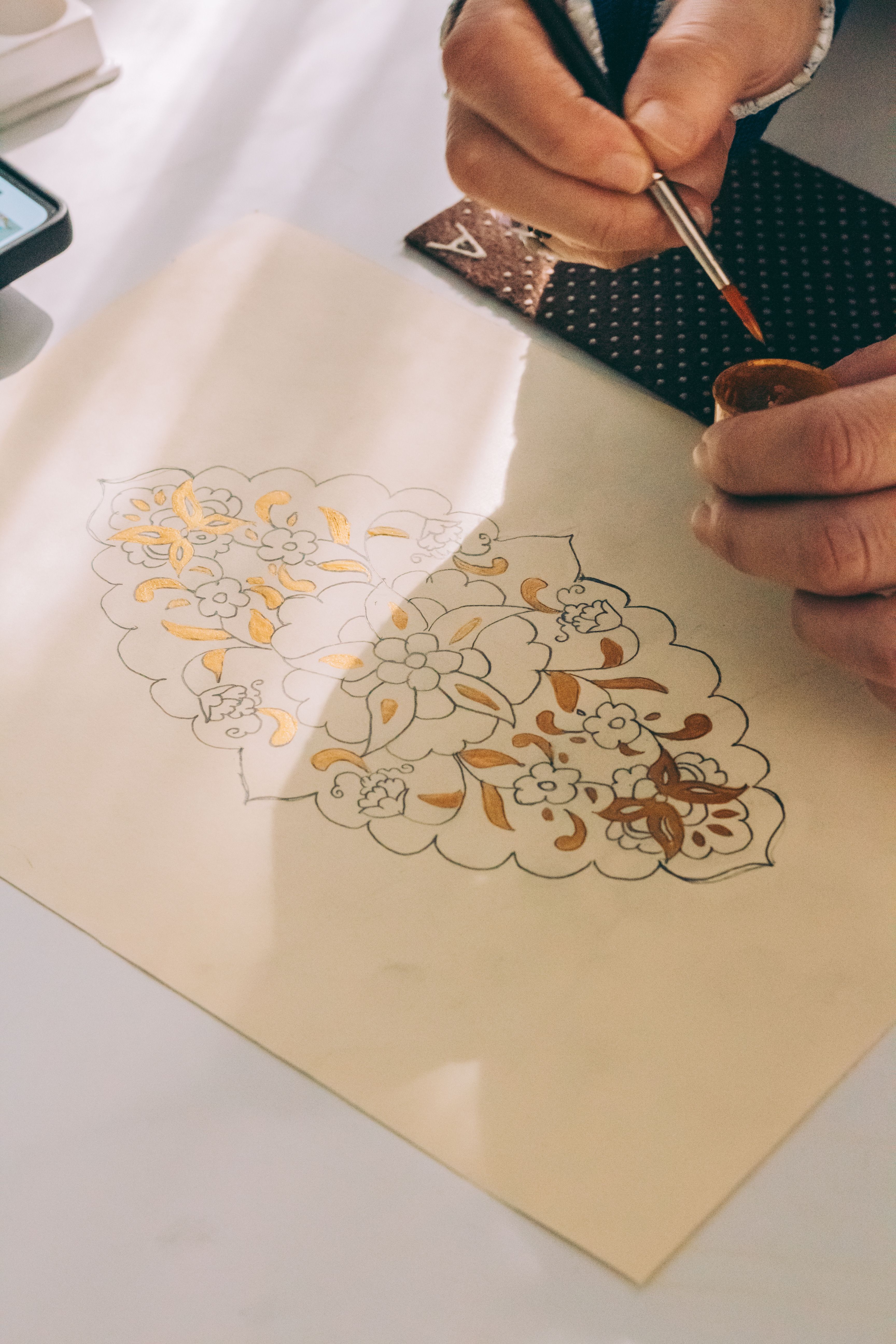 Close-up of a person painting a detailed floral design on paper using a brush and brown ink.