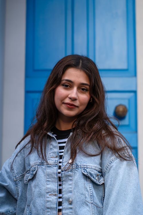 Free A young woman in a denim jacket stands confidently against a vibrant blue door. Stock Photo