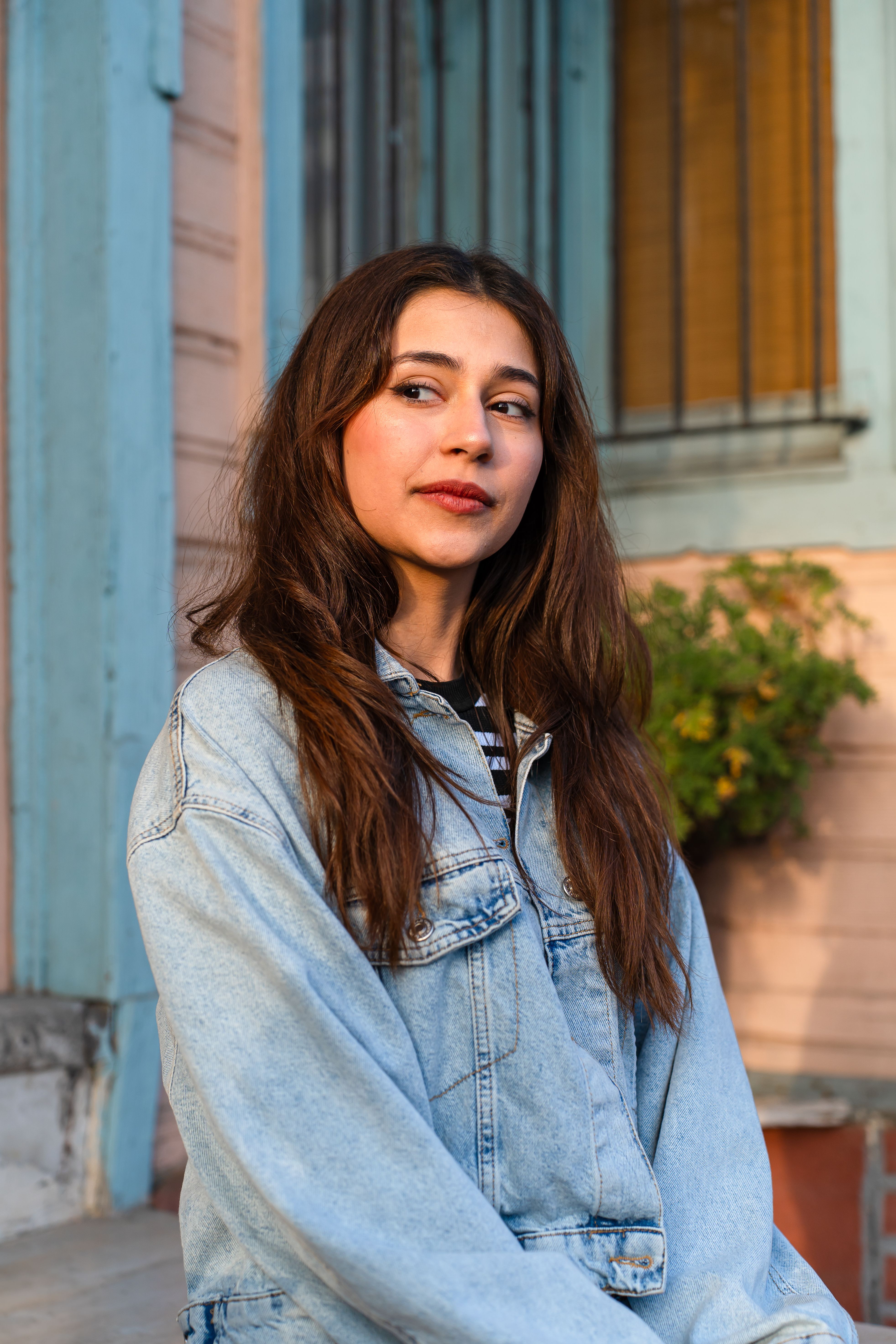 Portrait of a young woman in a denim jacket with a thoughtful expression, seated outside.