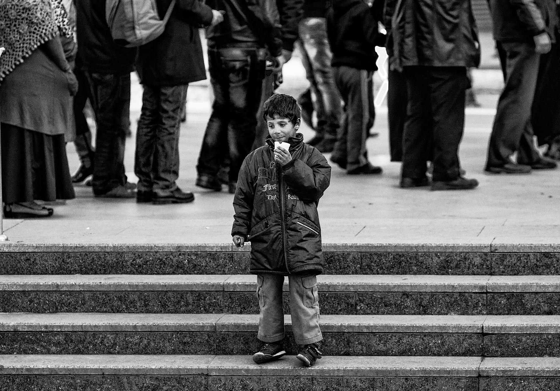 A solitary child in a coat stands on steps amidst a crowd, captured in a black and white scene.