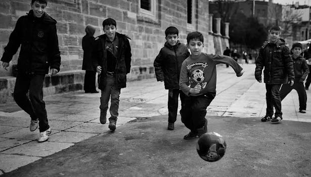 Black and white photo of children playing soccer, capturing dynamic motion and urban backdrop.