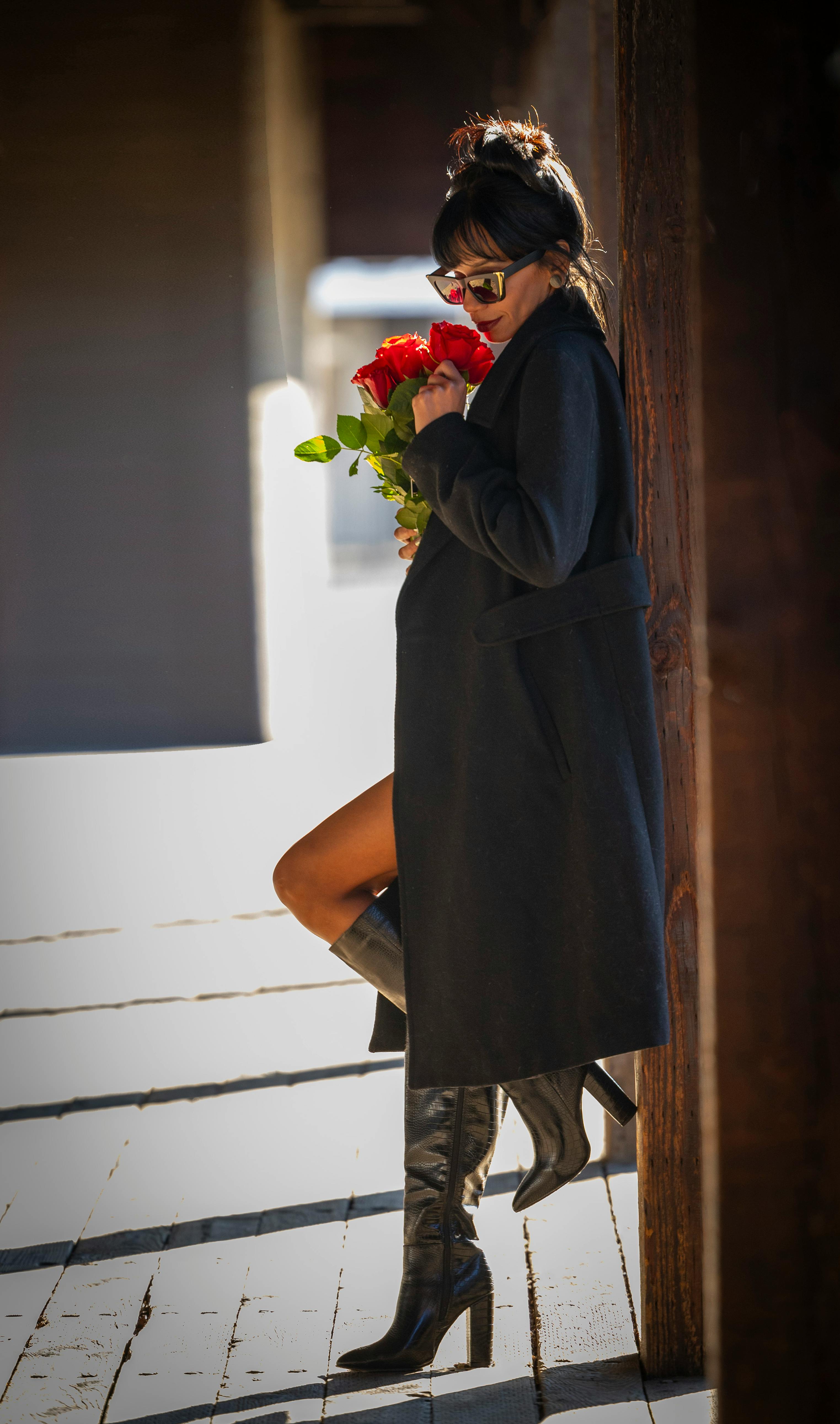 A fashionable woman in sunglasses and coat holds red roses, leaning against a wooden post outdoors.