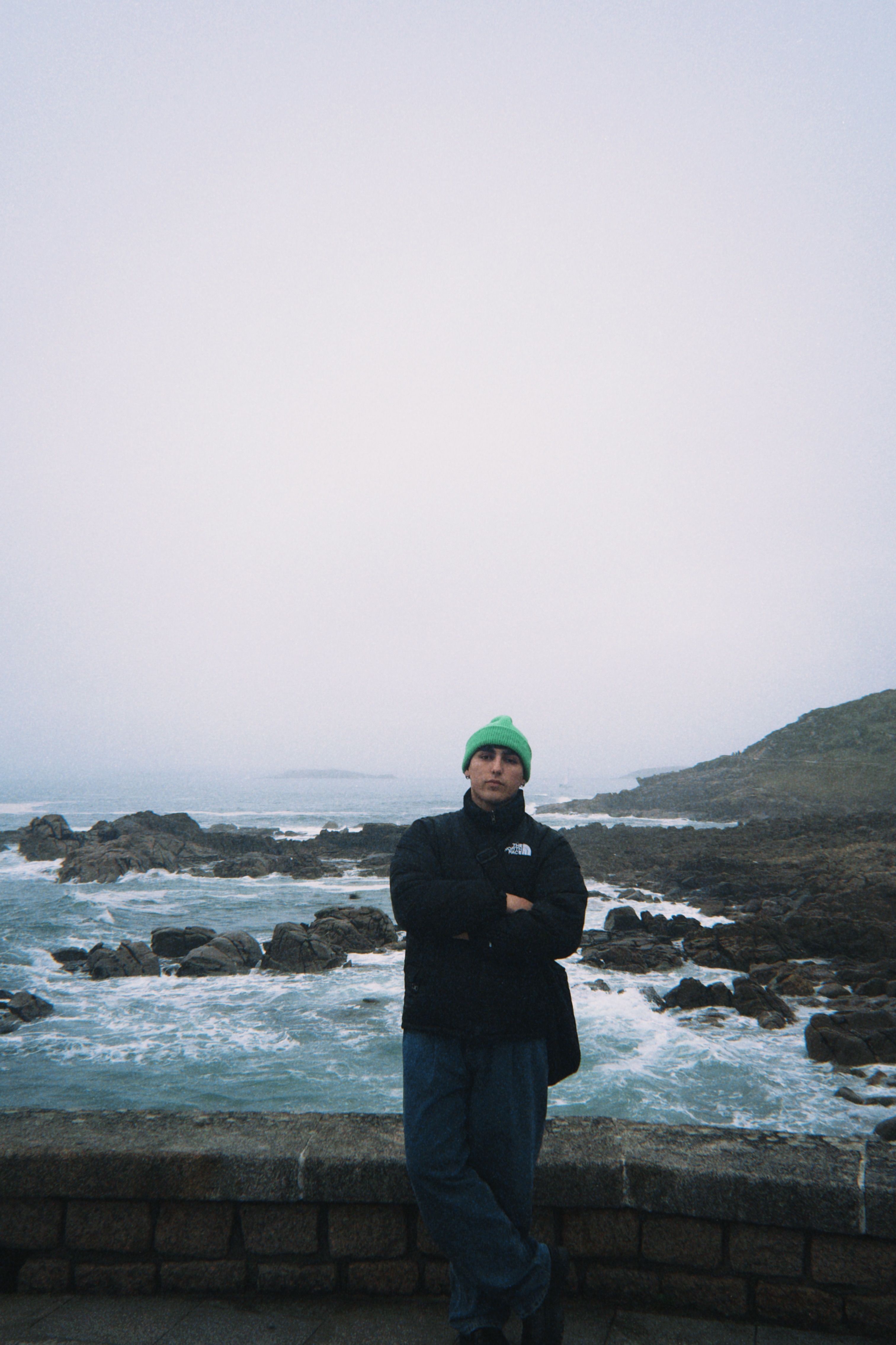 Man wearing green beanie standing by the ocean with rocky coastline.