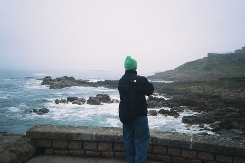 Free Person in green hat stands by the rocky ocean shore, embracing solitude and nature's beauty. Stock Photo