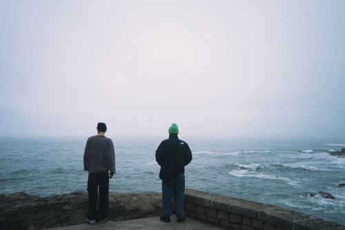 Free Two individuals stand on a rocky shore, looking out to a mist-covered ocean, evoking a sense of tranquility and introspection. Stock Photo