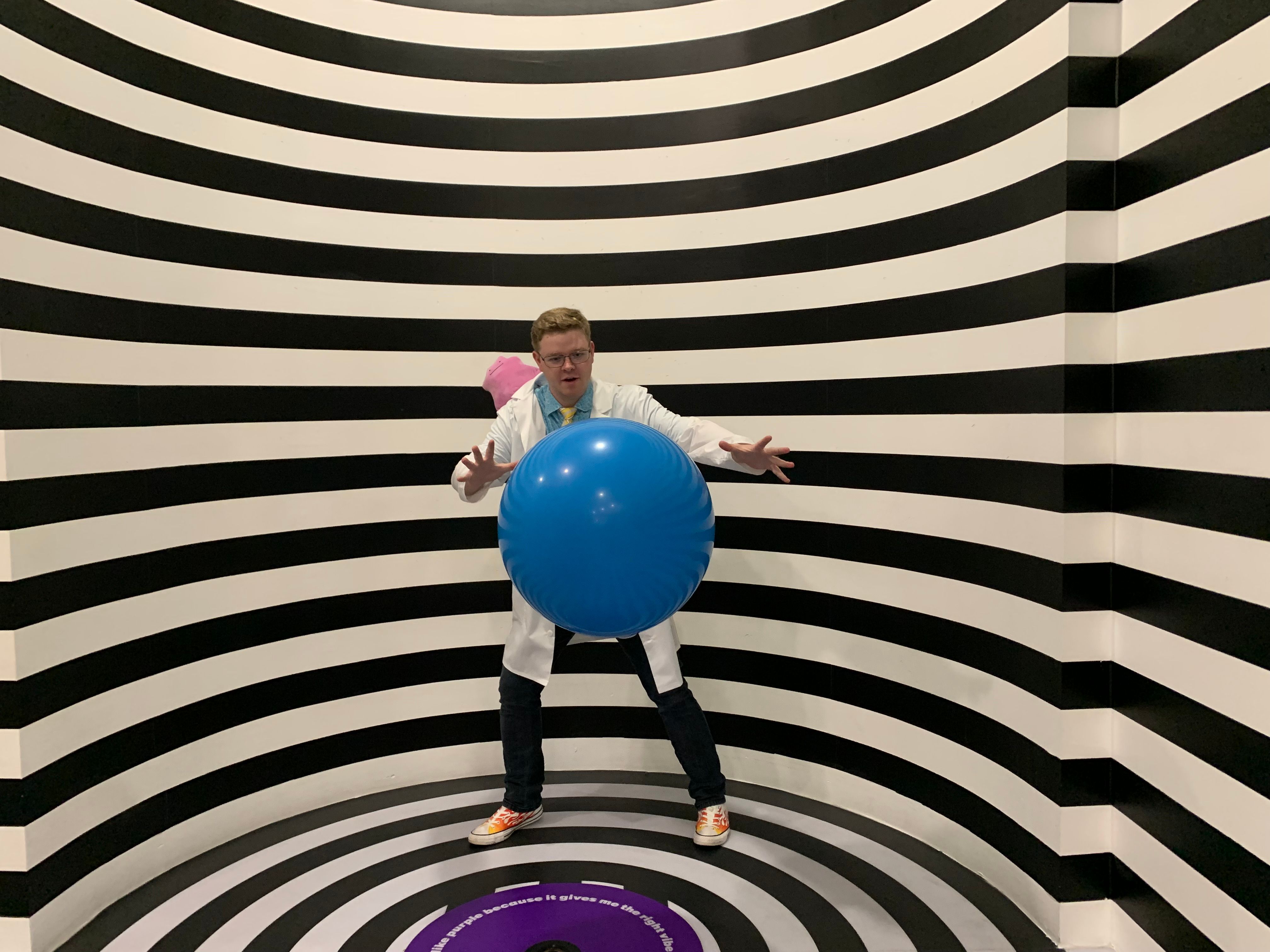 A man wearing a lab coat poses with a blue ball in a striped illusion room.