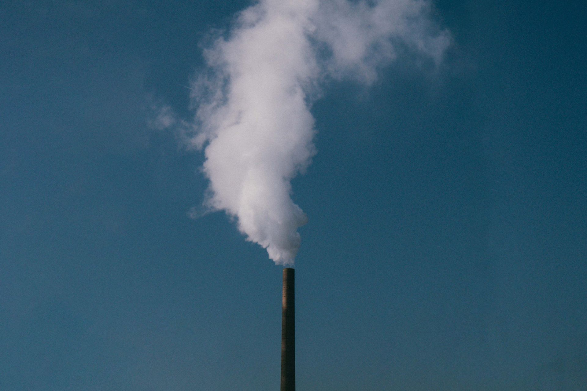 A factory smoke stack emitting white smoke under a clear blue sky, highlighting industrial impact on environment.