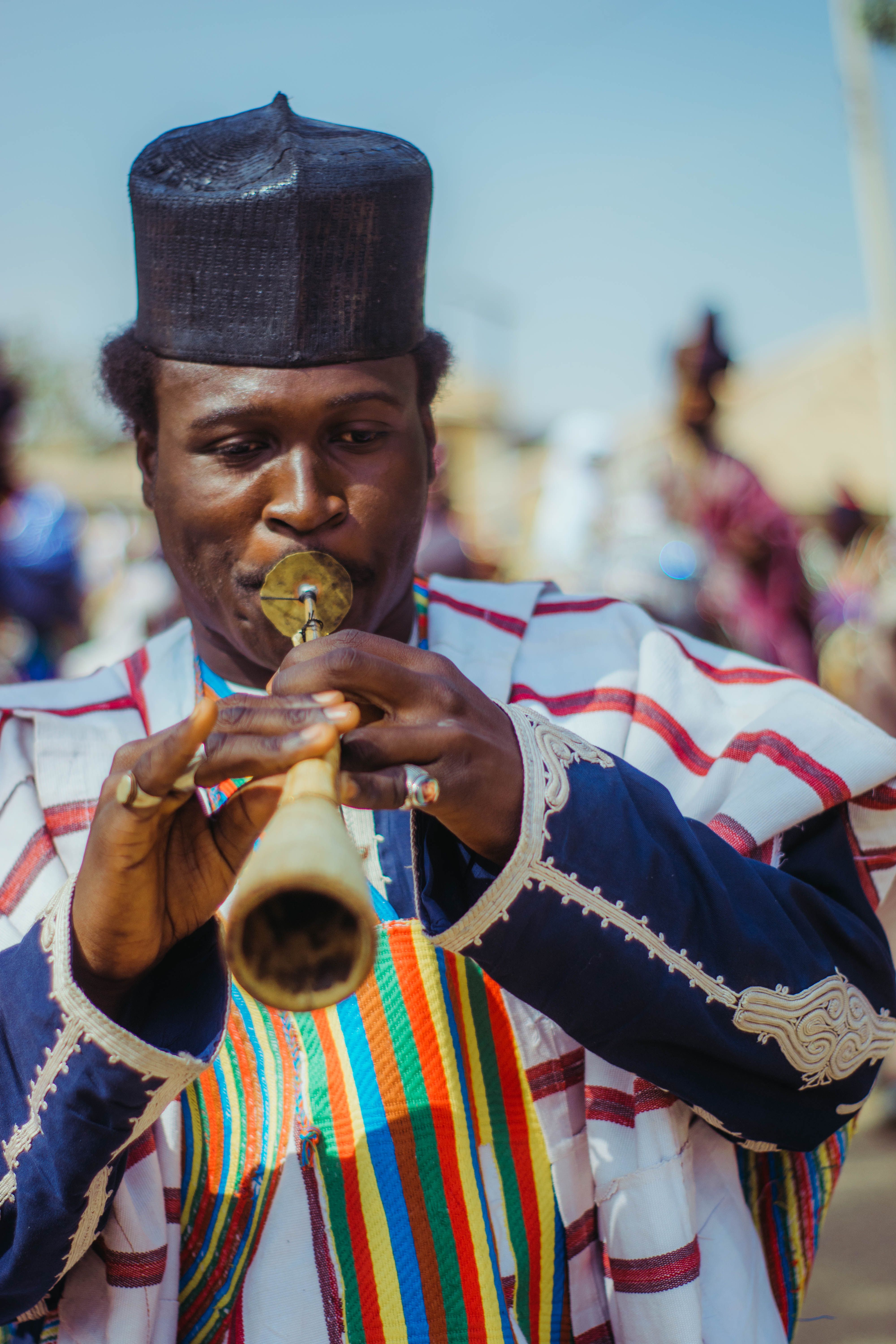 African musician in colorful attire playing a traditional wind instrument outdoors.