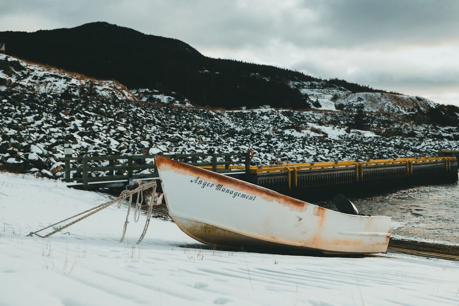 Rustic boat named 'Anger Management' resting on snowy shore by a rocky landscape.