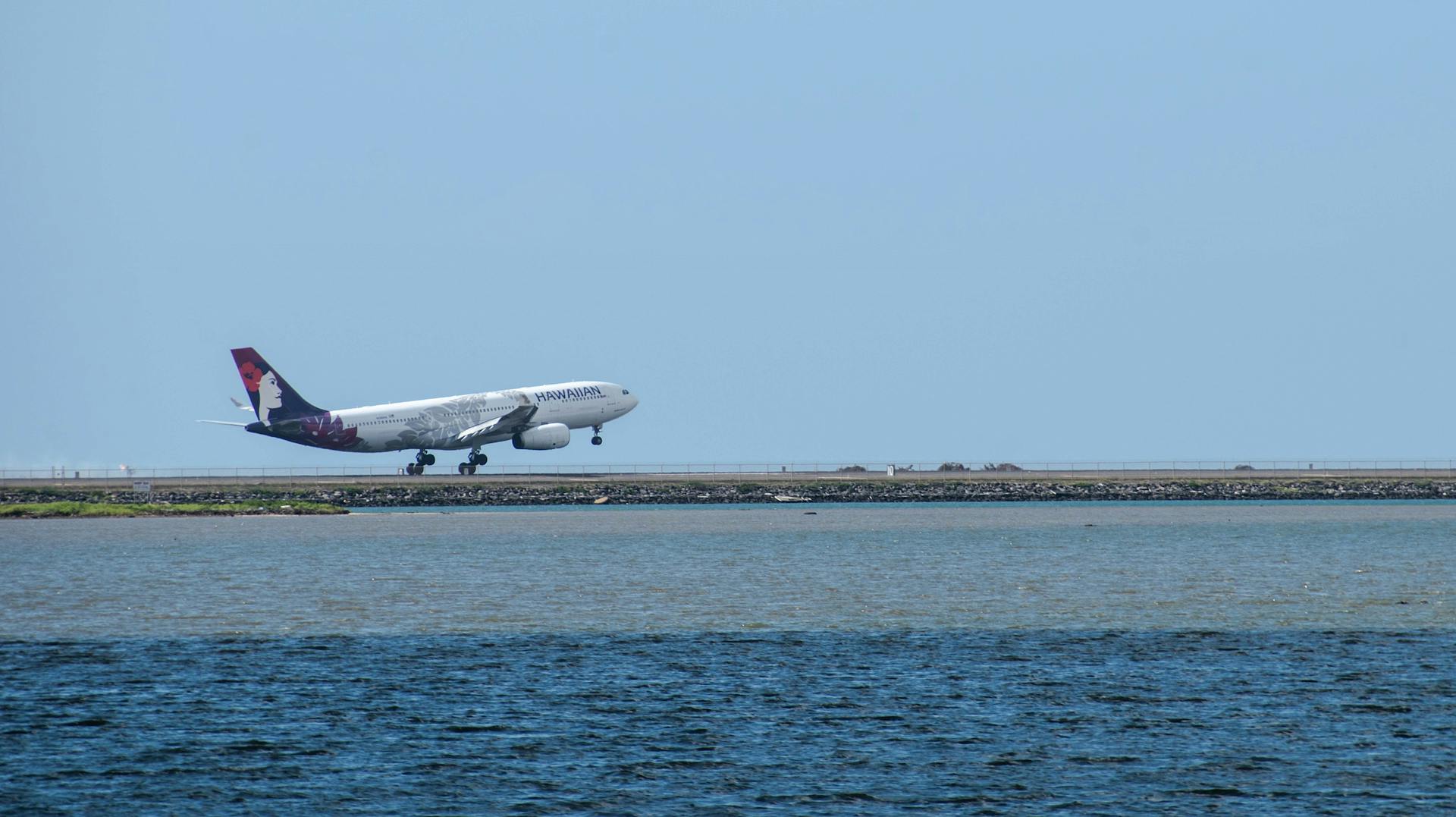 A Hawaiian Airlines plane taking off from a runway near the ocean on a clear day.