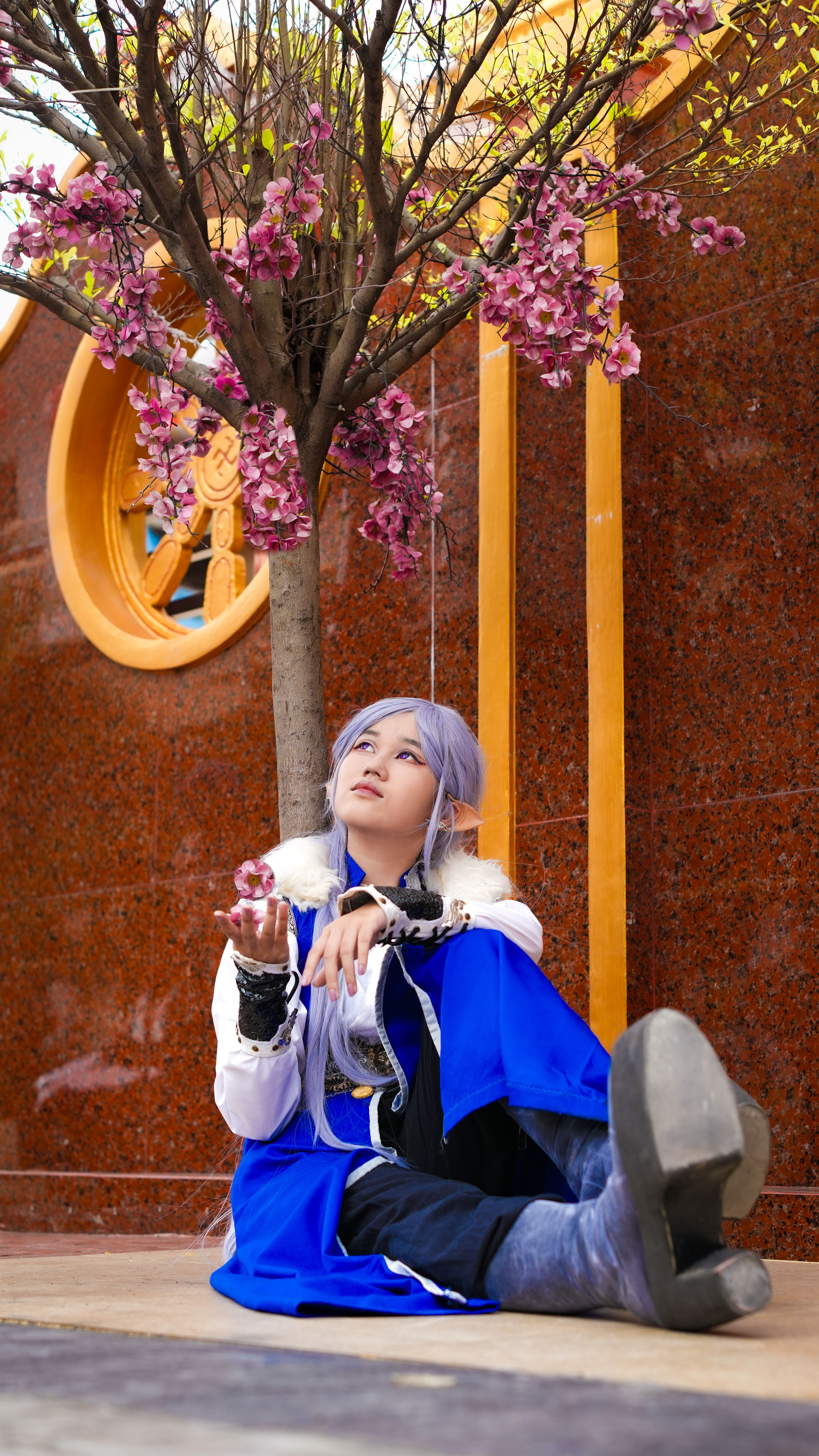 A young Asian model in a blue cosplay outfit posing under cherry blossoms, showcasing a serene expression.