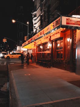 Nighttime cityscape of a lively New York City street with a glowing restaurant and a classic yellow taxi.