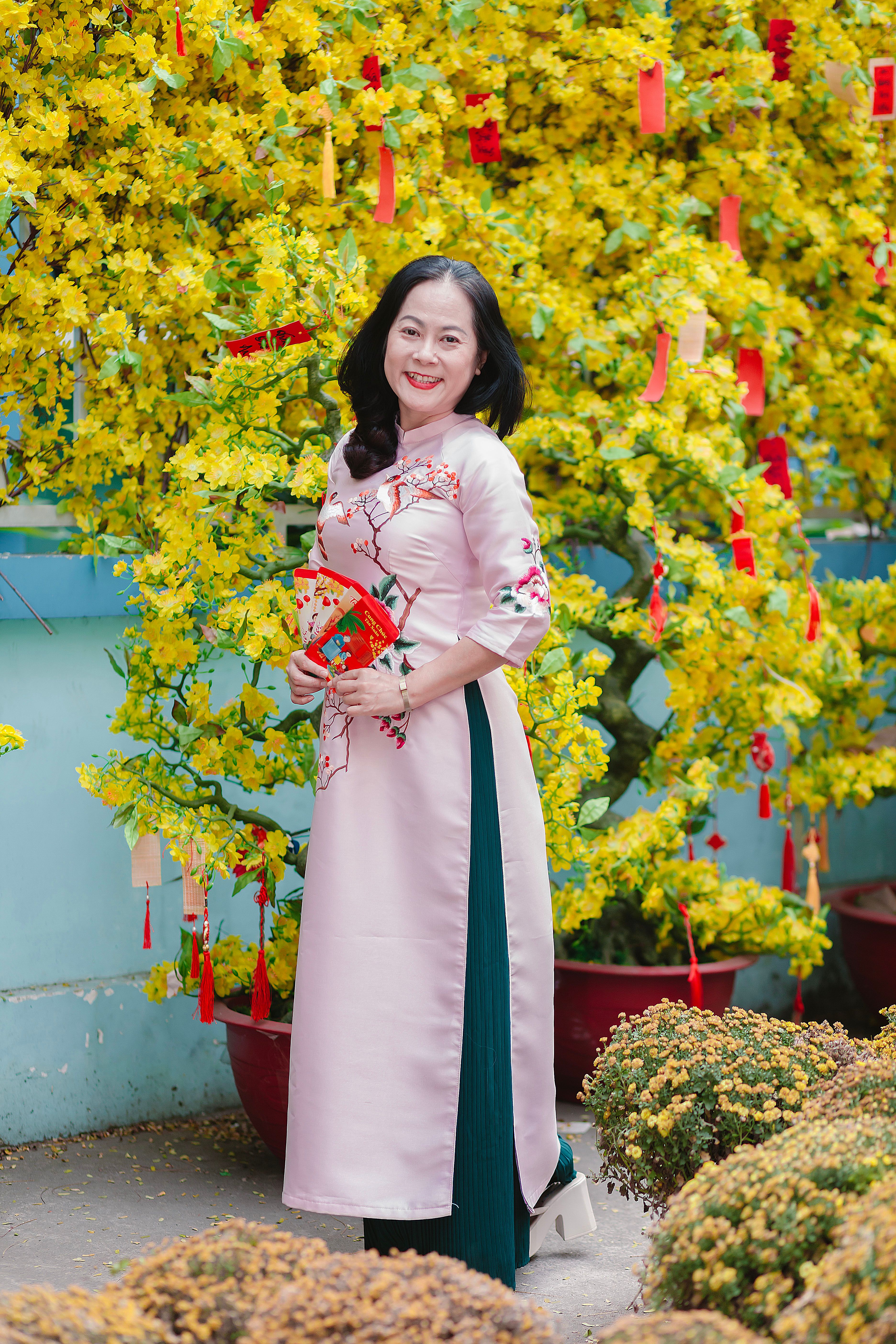 Woman in traditional dress celebrating Tet with yellow flowers and red decorations outdoors.