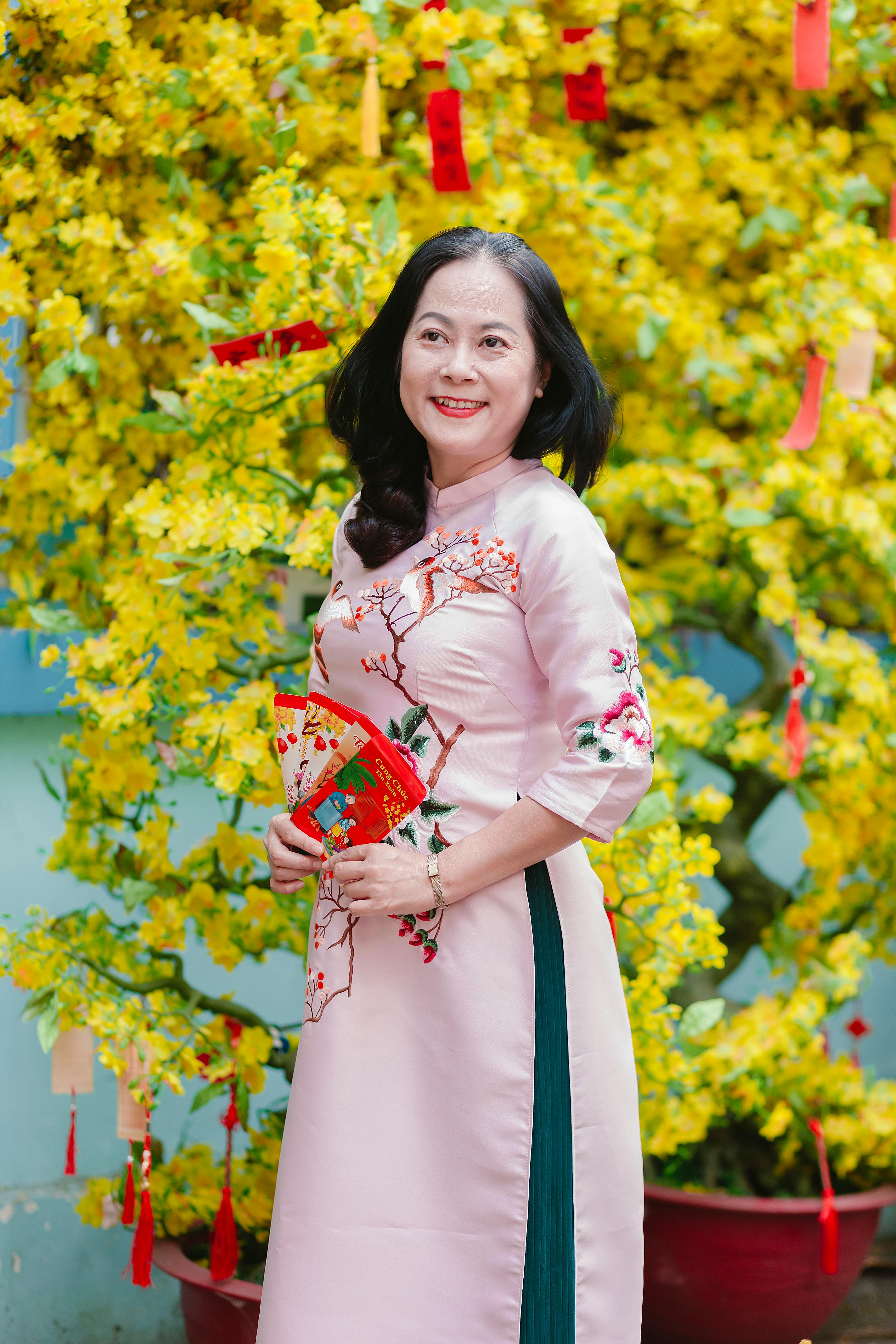 Smiling woman in traditional dress holding envelopes in front of a blooming yellow tree.