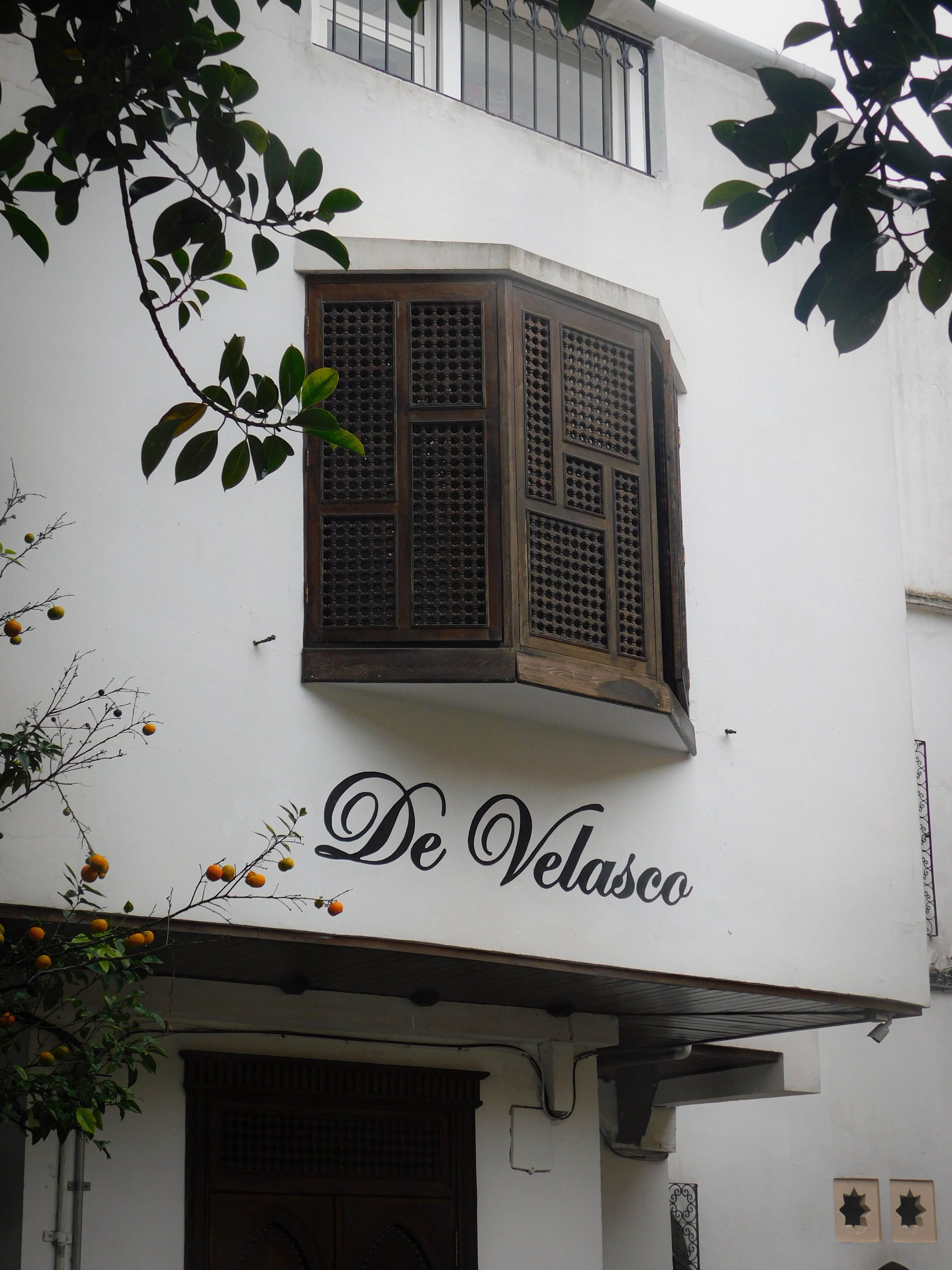 A classic bay window with wooden shutters on a white building exterior with 'De Velasco' signage.