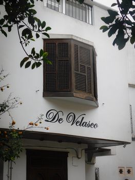 A classic bay window with wooden shutters on a white building exterior with 'De Velasco' signage.