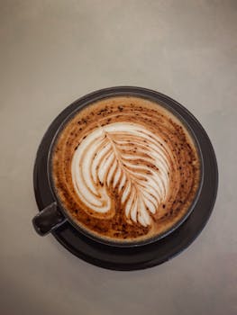 Latte with intricate leaf foam art in a dark cup on a neutral background, perfect for cafe aesthetics.
