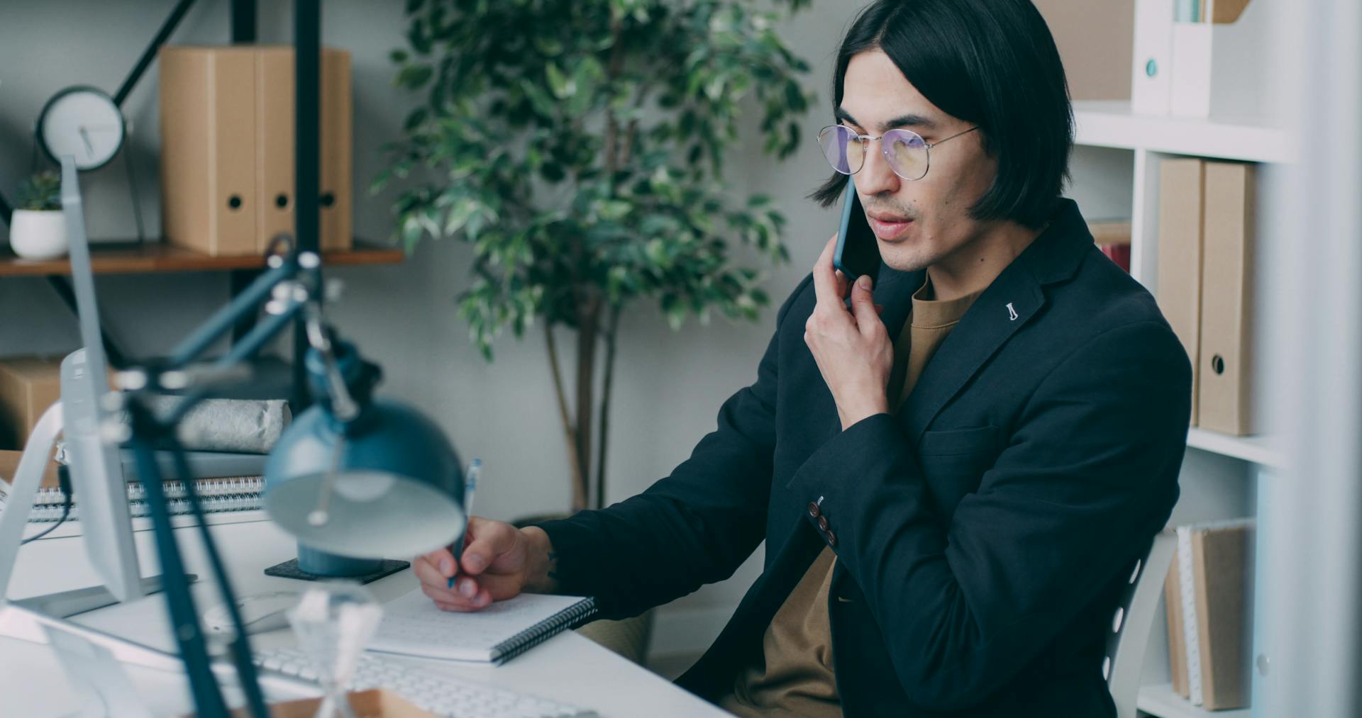 A man in glasses is multitasking by talking on the phone and writing notes in a modern office setting.