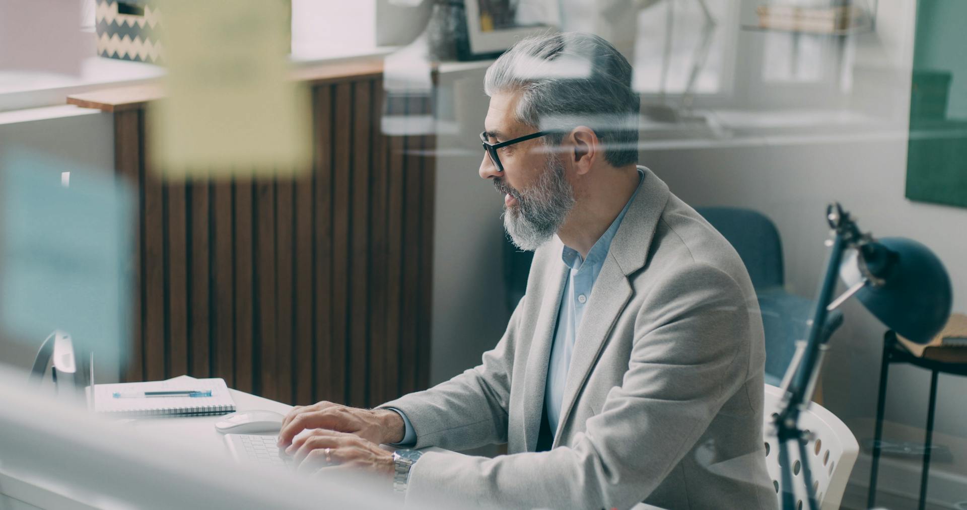 A senior man in a gray suit working at his desk in a modern office setting.