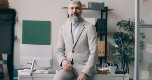 Free A mature businessman in a beige suit sits confidently on a desk in a modern office setting. Stock Photo