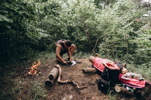 Man setting up campfire in forest with gear spread out. Perfect for outdoor adventure themes.