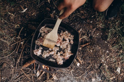 Person grilling meat outdoors over an open fire, showing rustic camp cooking.