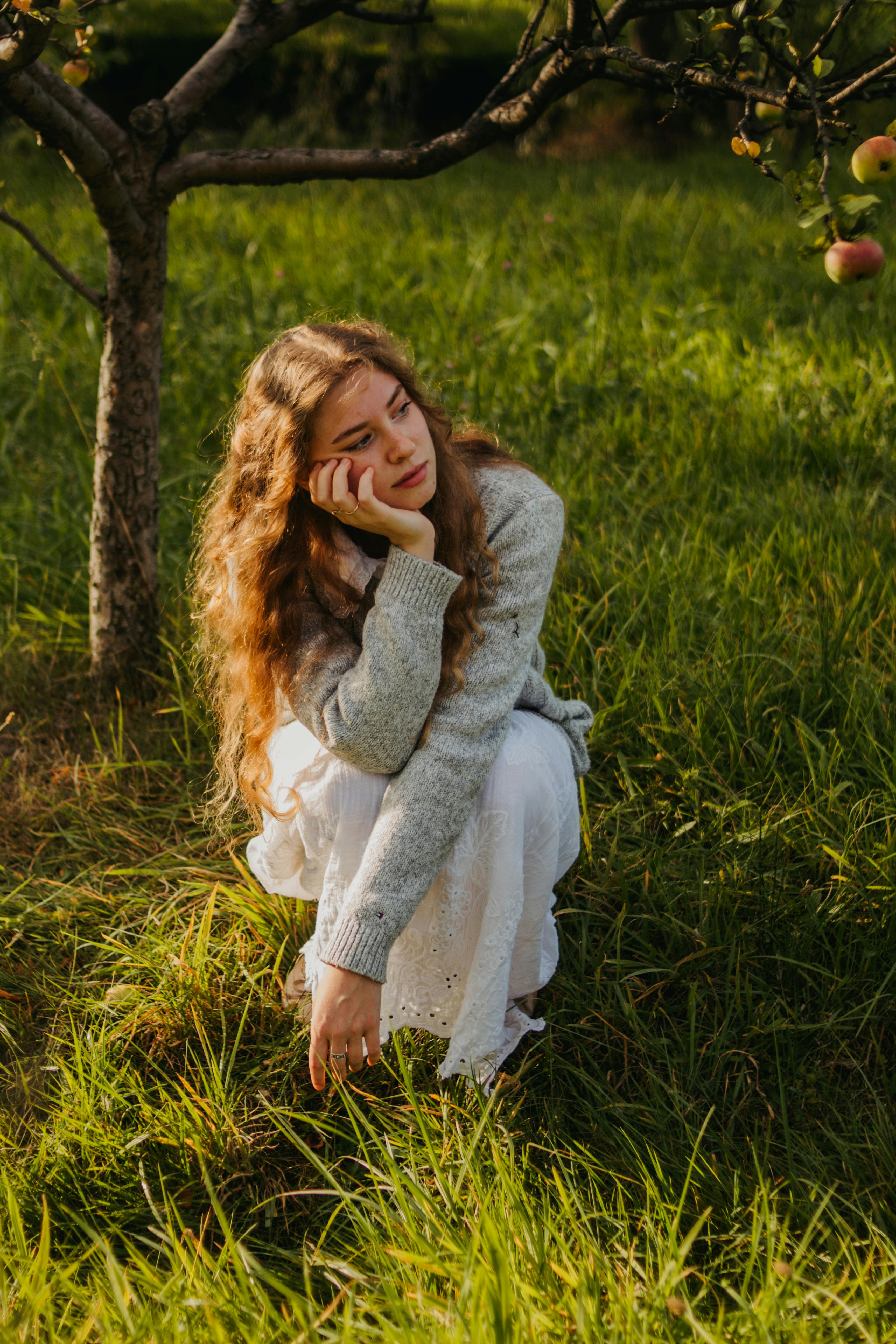Young woman with long hair sitting pensively in a sunlit apple orchard.