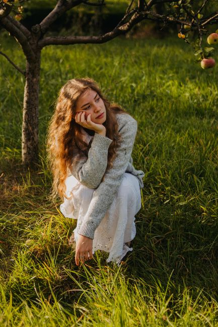 Young woman with long hair sitting pensively in a sunlit apple orchard.
