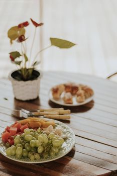 A rustic fruit platter with grapes and melon on a wooden table with bread rolls in the background.