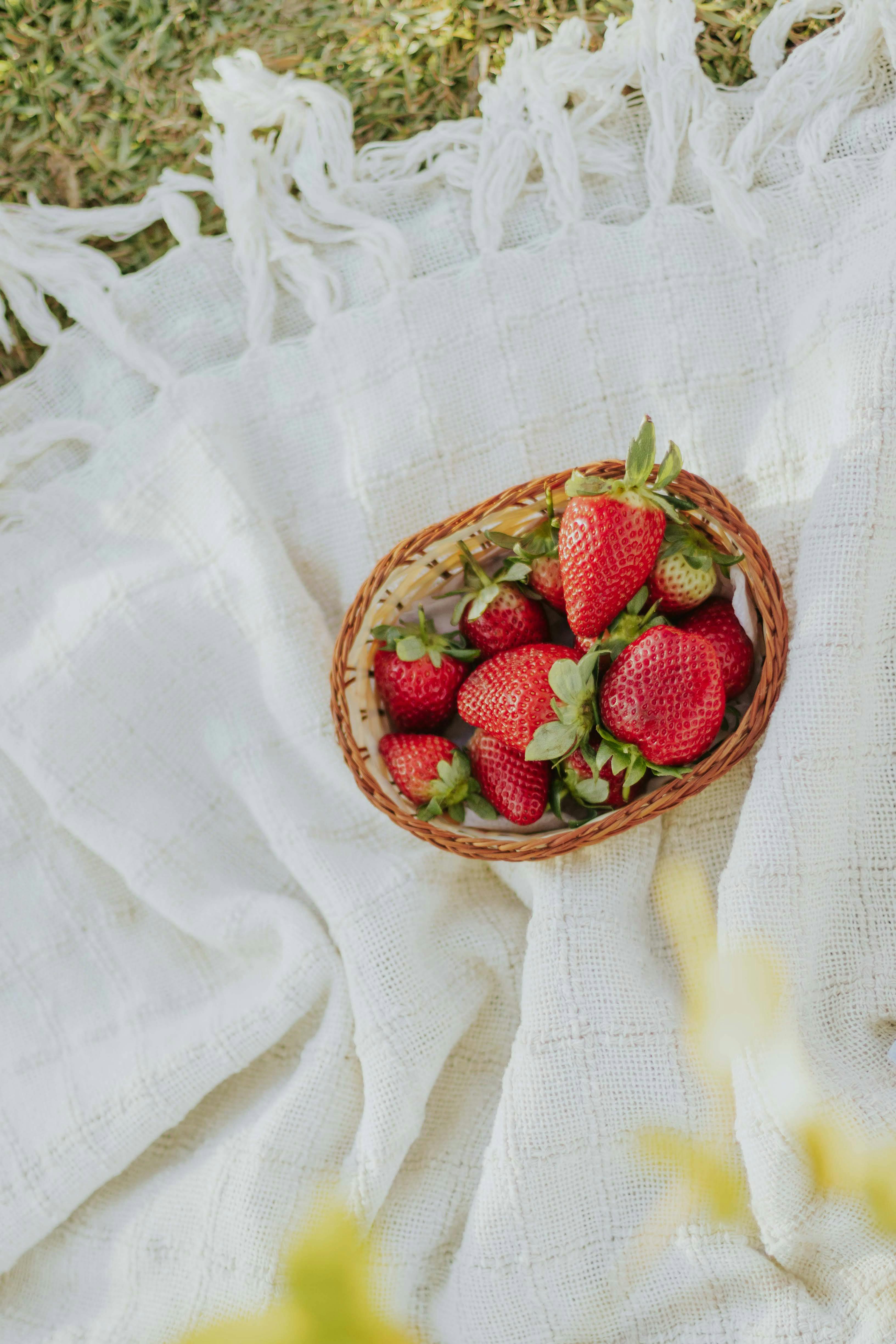 Wicker basket filled with ripe strawberries placed on a white blanket in natural light.
