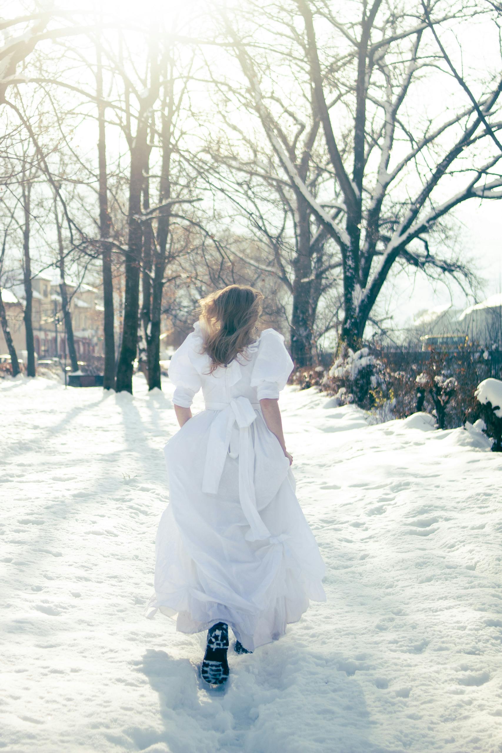 A woman in a white dress walks through a snowy landscape on a bright winter day.