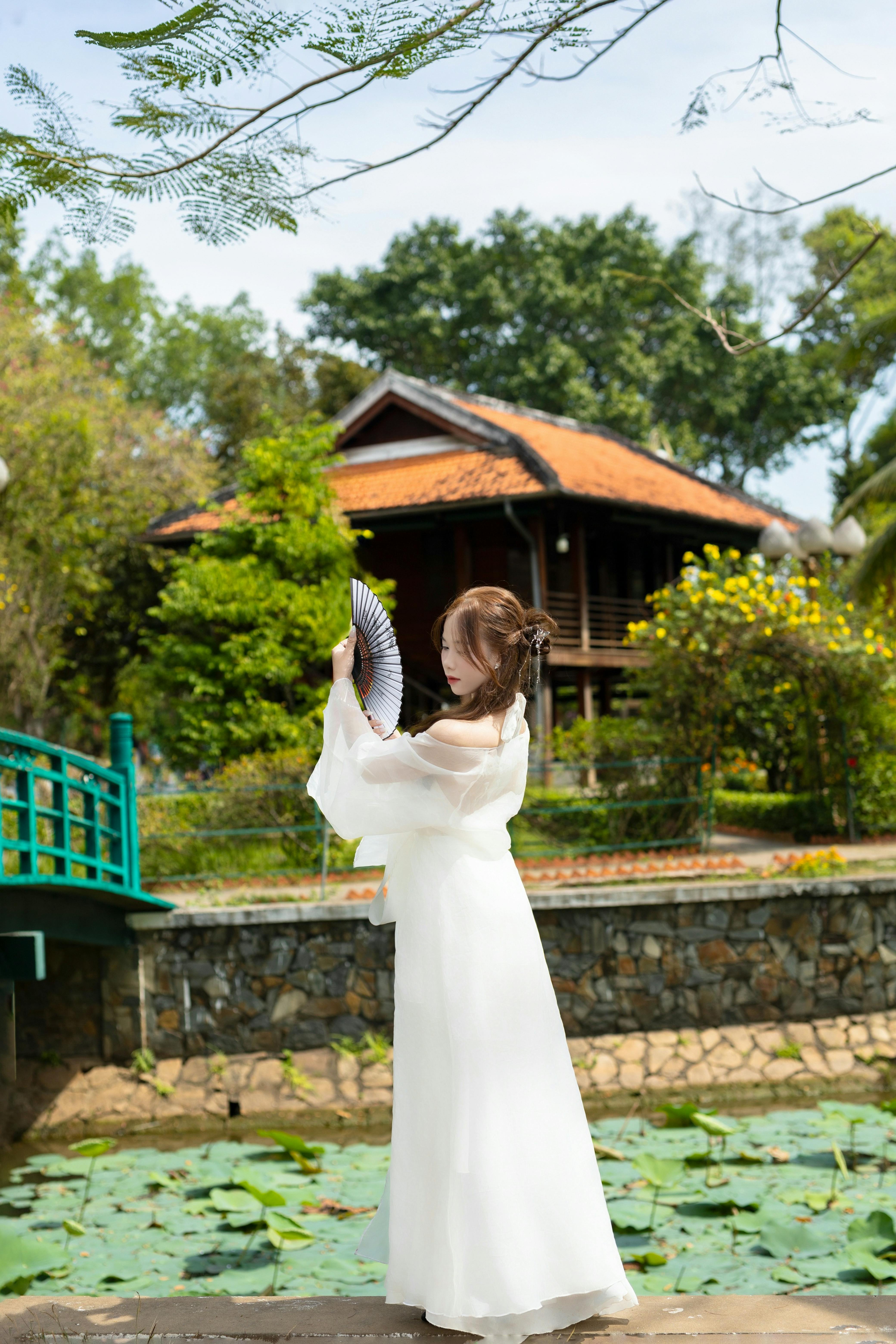 Woman in traditional attire holding fan near rustic Asian house.